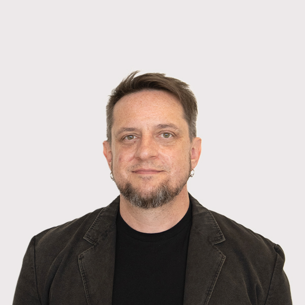 Professional portrait of a man with short hair, wearing a black shirt and a dark blazer, set against a plain white background.
